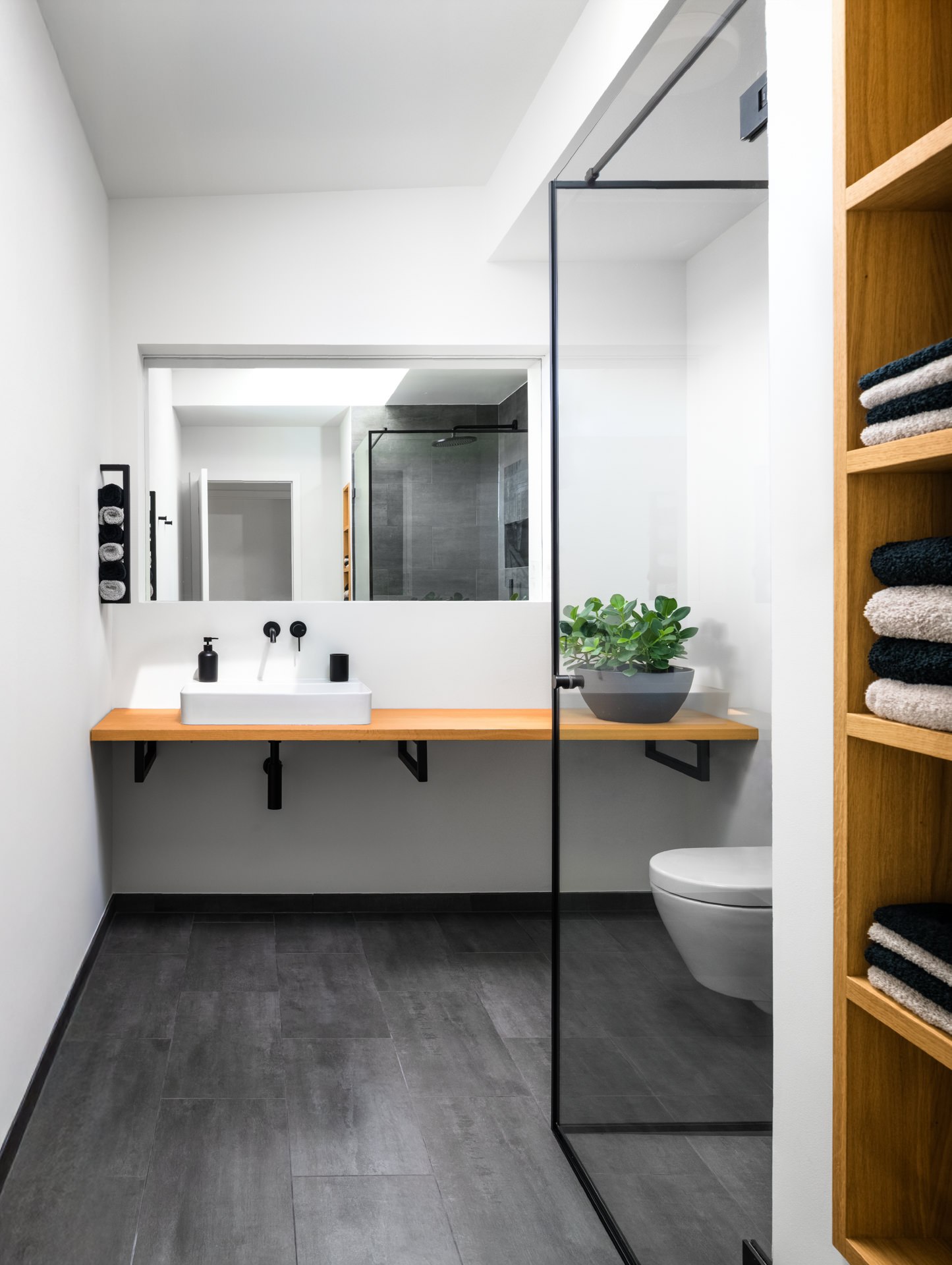 Minimalist black and white bathroom with floating wooden vanity, dark tile floor, and glass shower partition