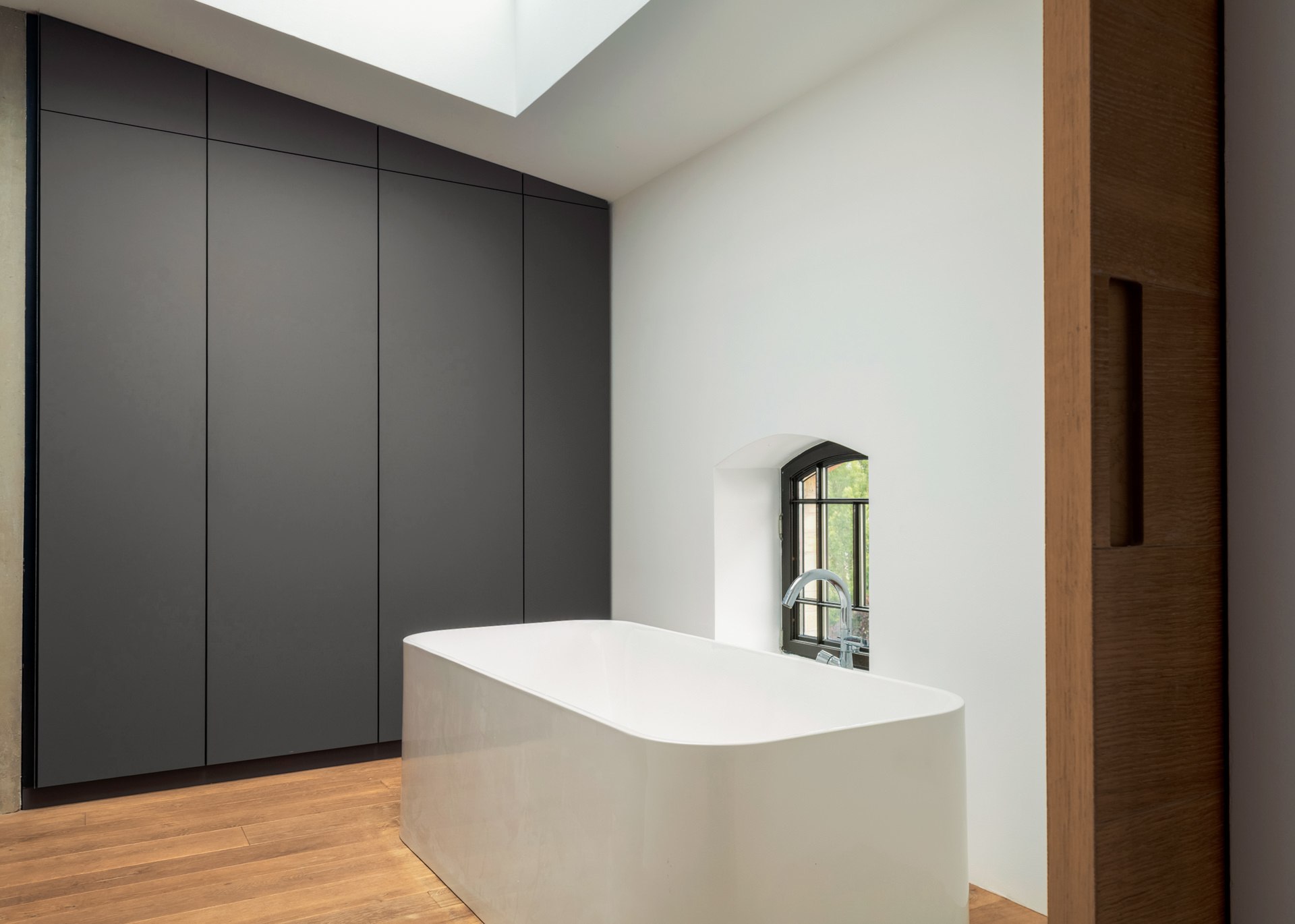 Bright master bathroom with a freestanding white bathtub beside a tall arched window — architectural interior photography