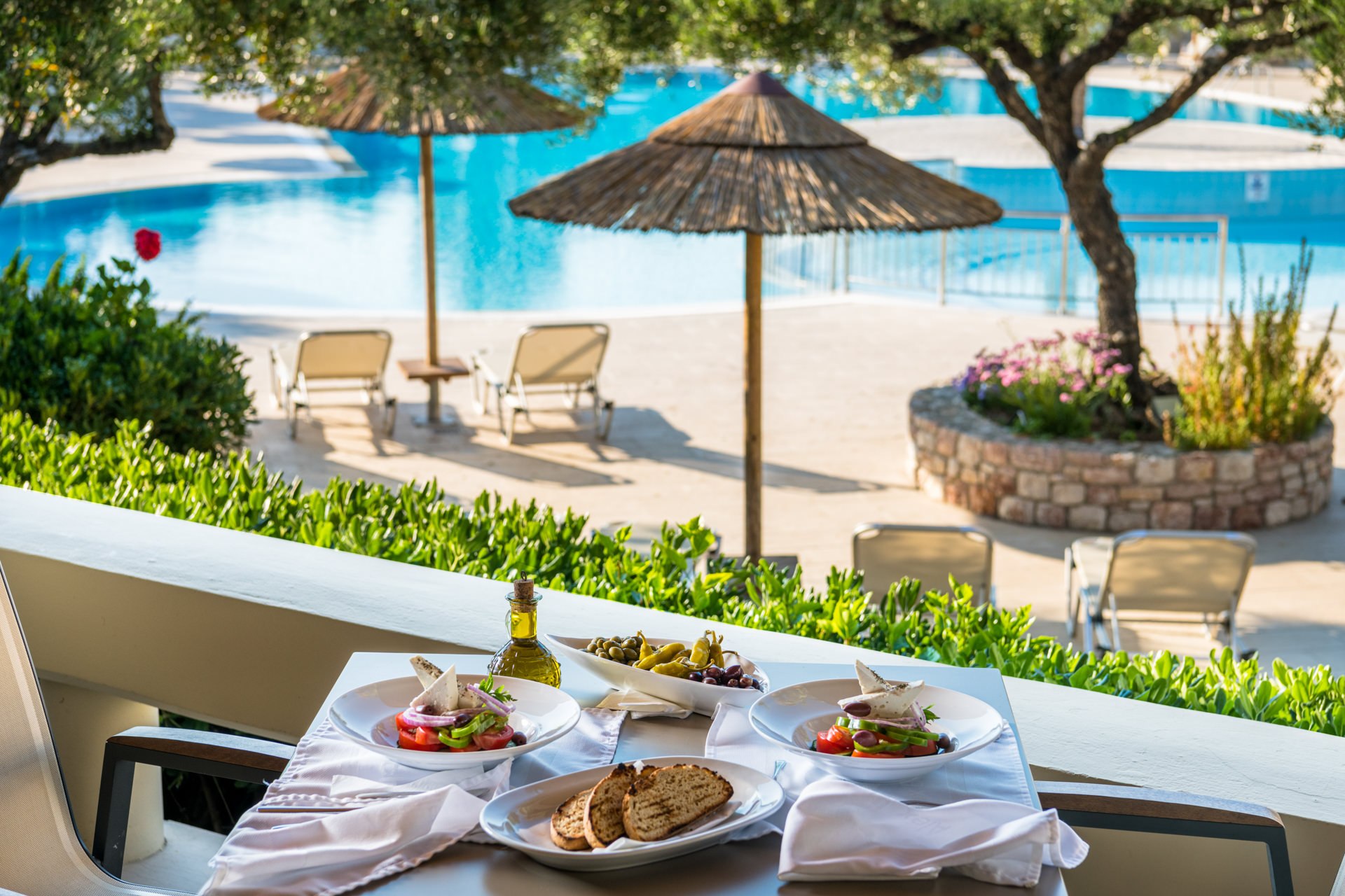 Elegantly set outdoor buffet restaurant table with white cloth and food, overlooking a hotel pool with thatched parasols