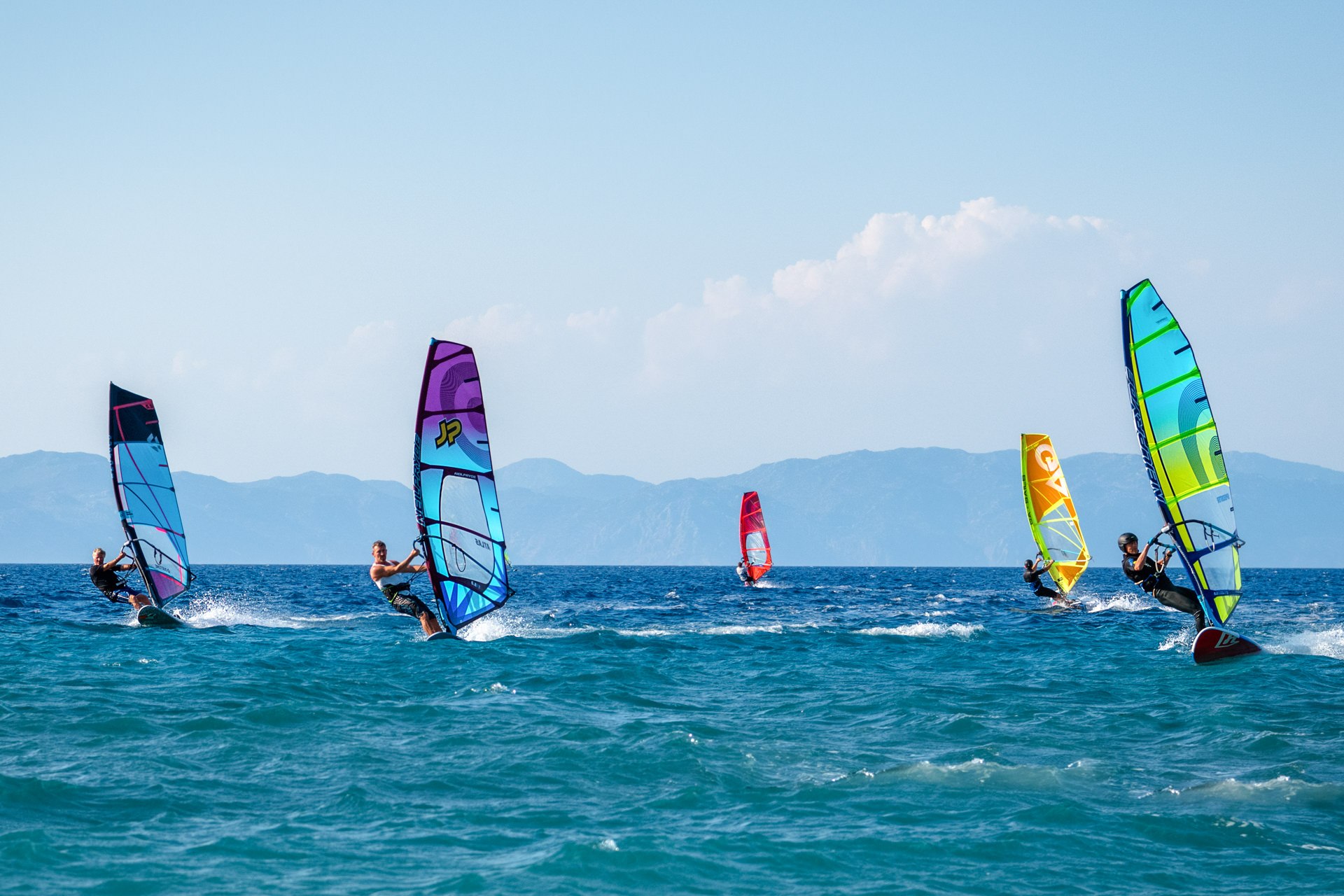 Multiple colorful windsurfers on a bright blue sea with mountains in the background — water sports destination photography