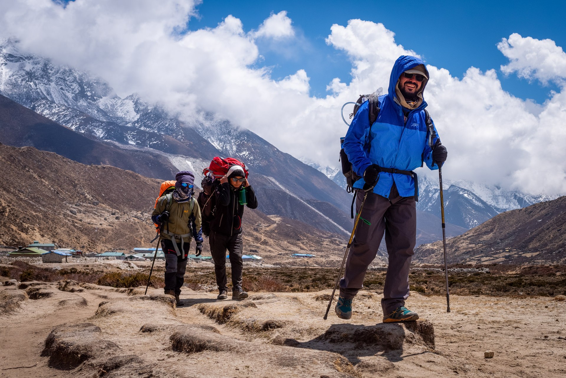 hree trekkers in colorful gear hiking through a high-altitude Himalayan valley with snow-covered peaks and clouds
