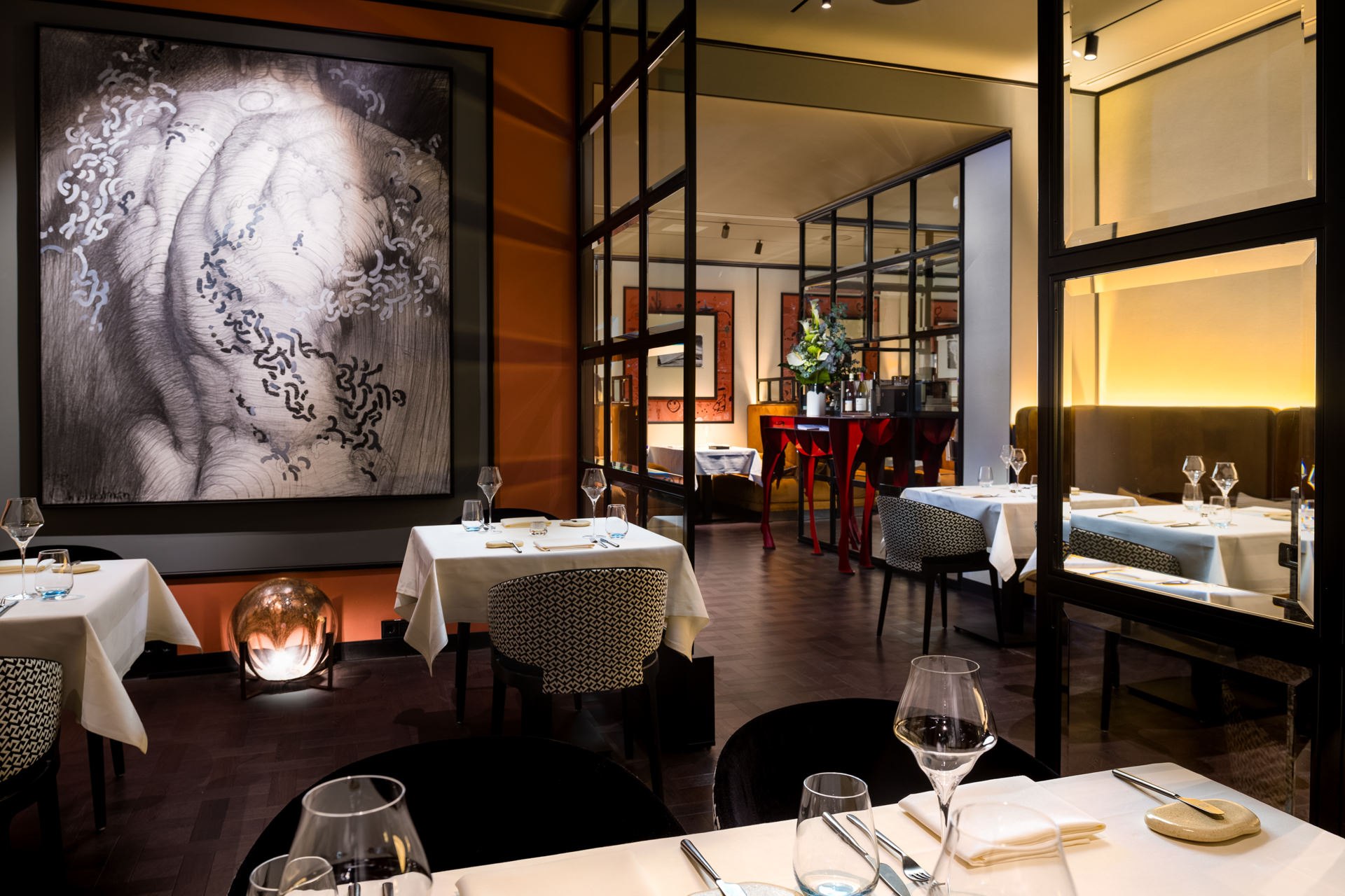 Sophisticated restaurant dining room with large-scale artwork, dark steel-frame glass partition, and white linen settings