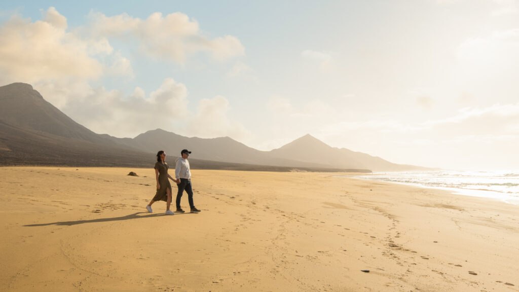 Two people walking on the immense empty beach of Cofete, Fuerteventura, with volcanic mountains in the background