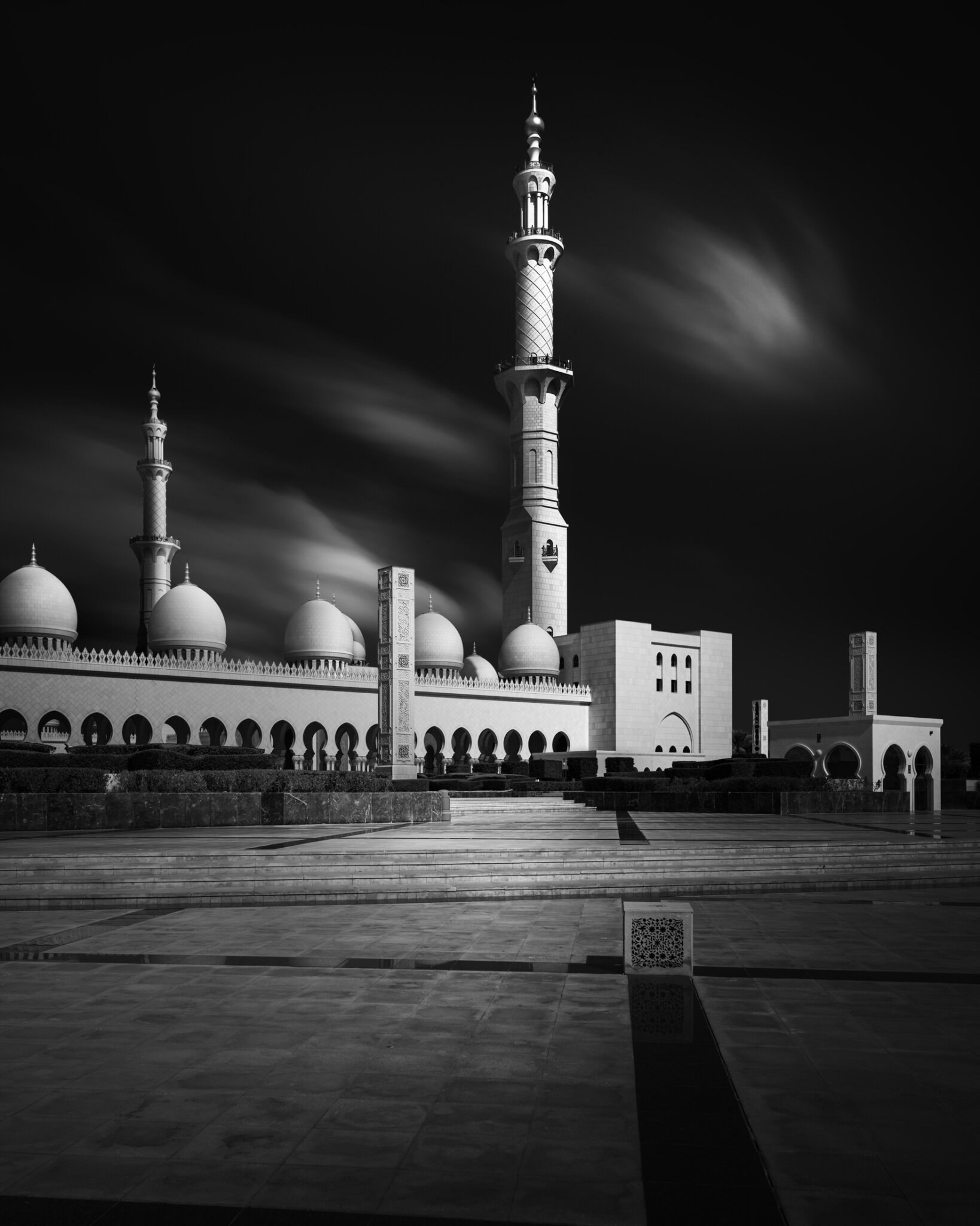 Long exposure black and white fine art photograph of a grand mosque with a tall minaret and dramatically streaking clouds
