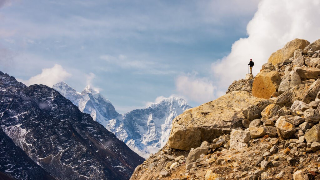 Lone trekker standing on a rocky high-altitude ridge surrounded by mist and towering Himalayan peaks