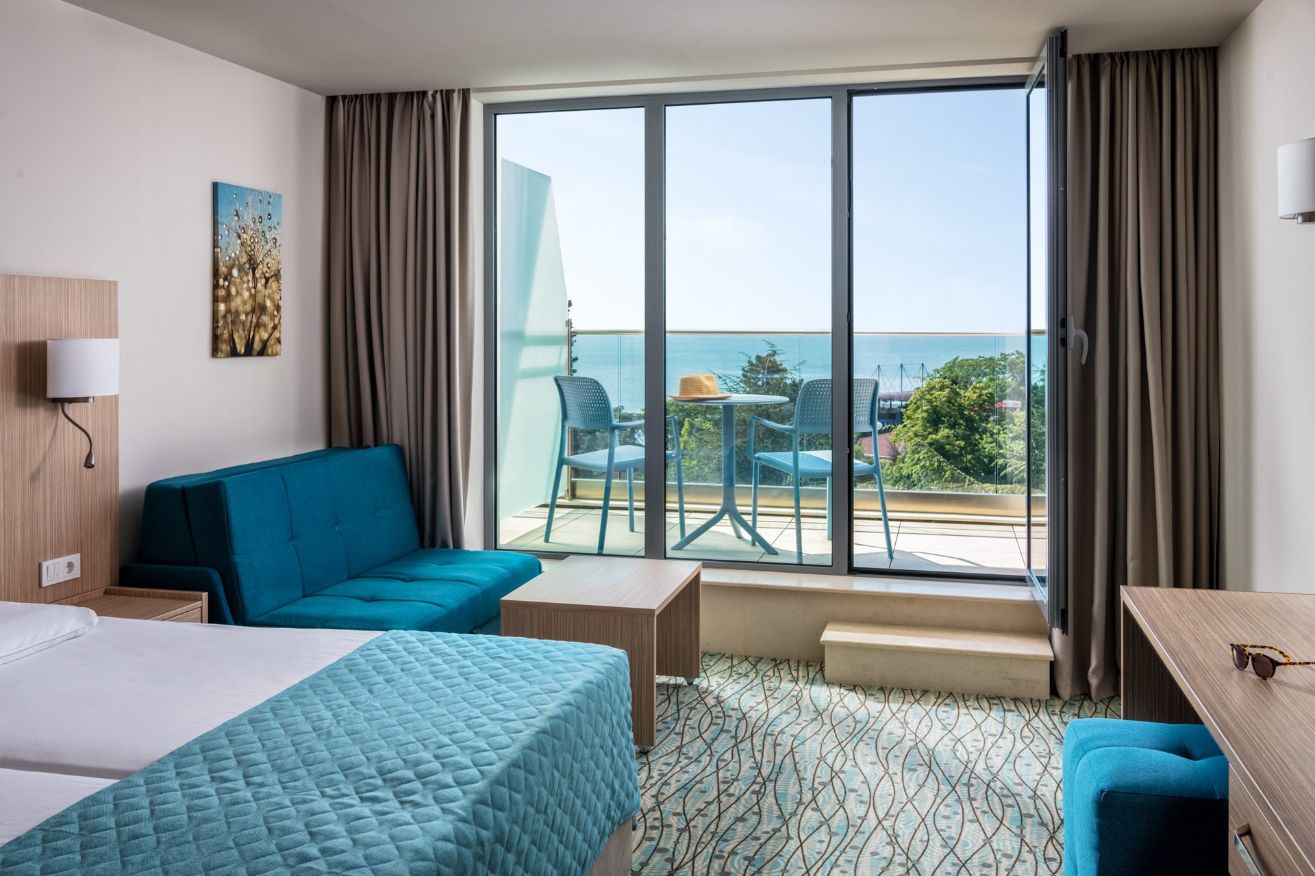 Bright hotel room with floor-to-ceiling glass doors, ocean view, blue accent sofa, and private balcony