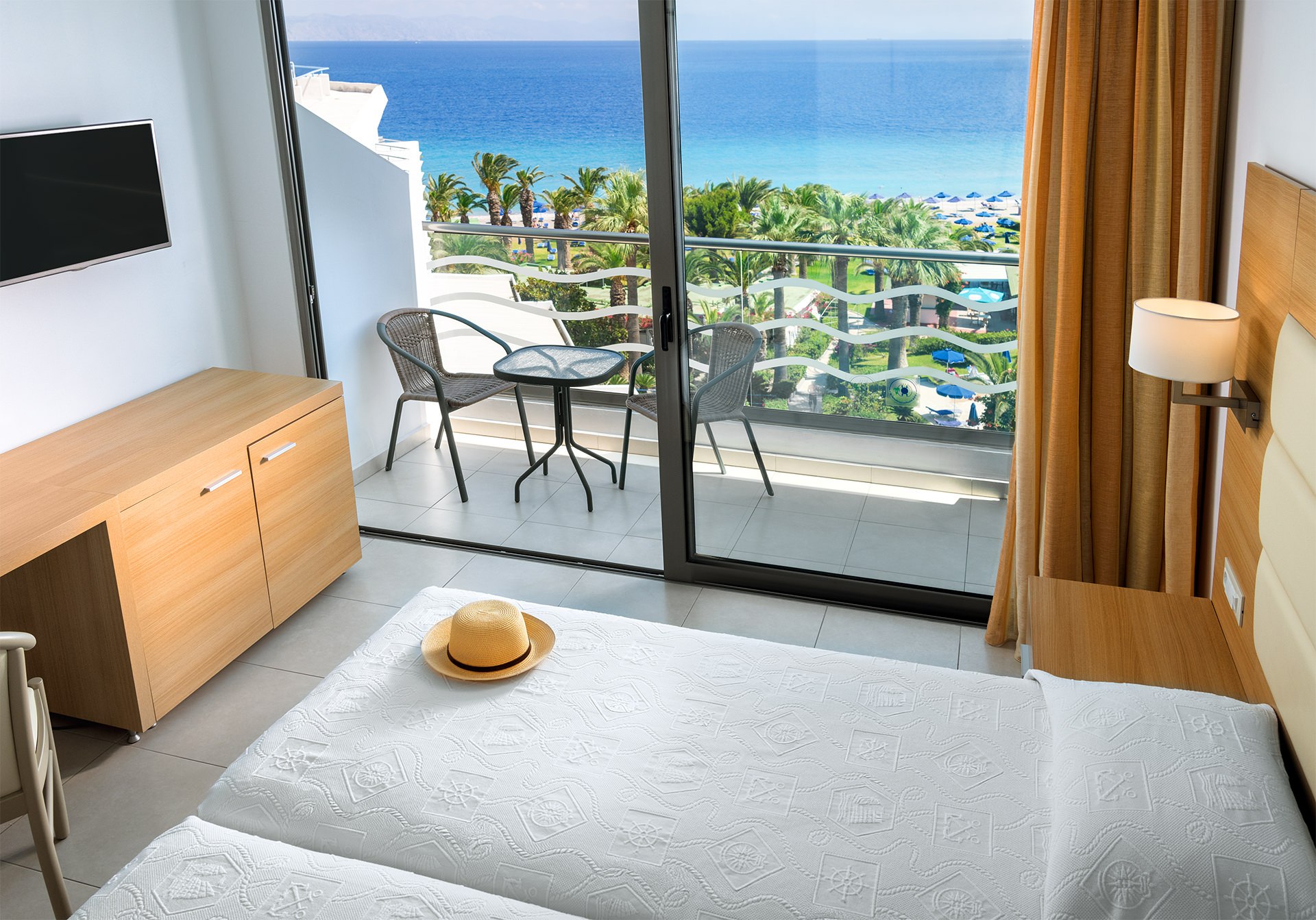 Modern hotel room with a bed, wooden furniture, and a balcony overlooking the ocean and palm trees