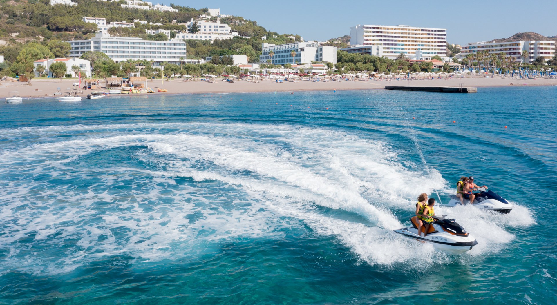 Aerial photograph of a jet ski creating a white wake along a clear blue Mediterranean coastline