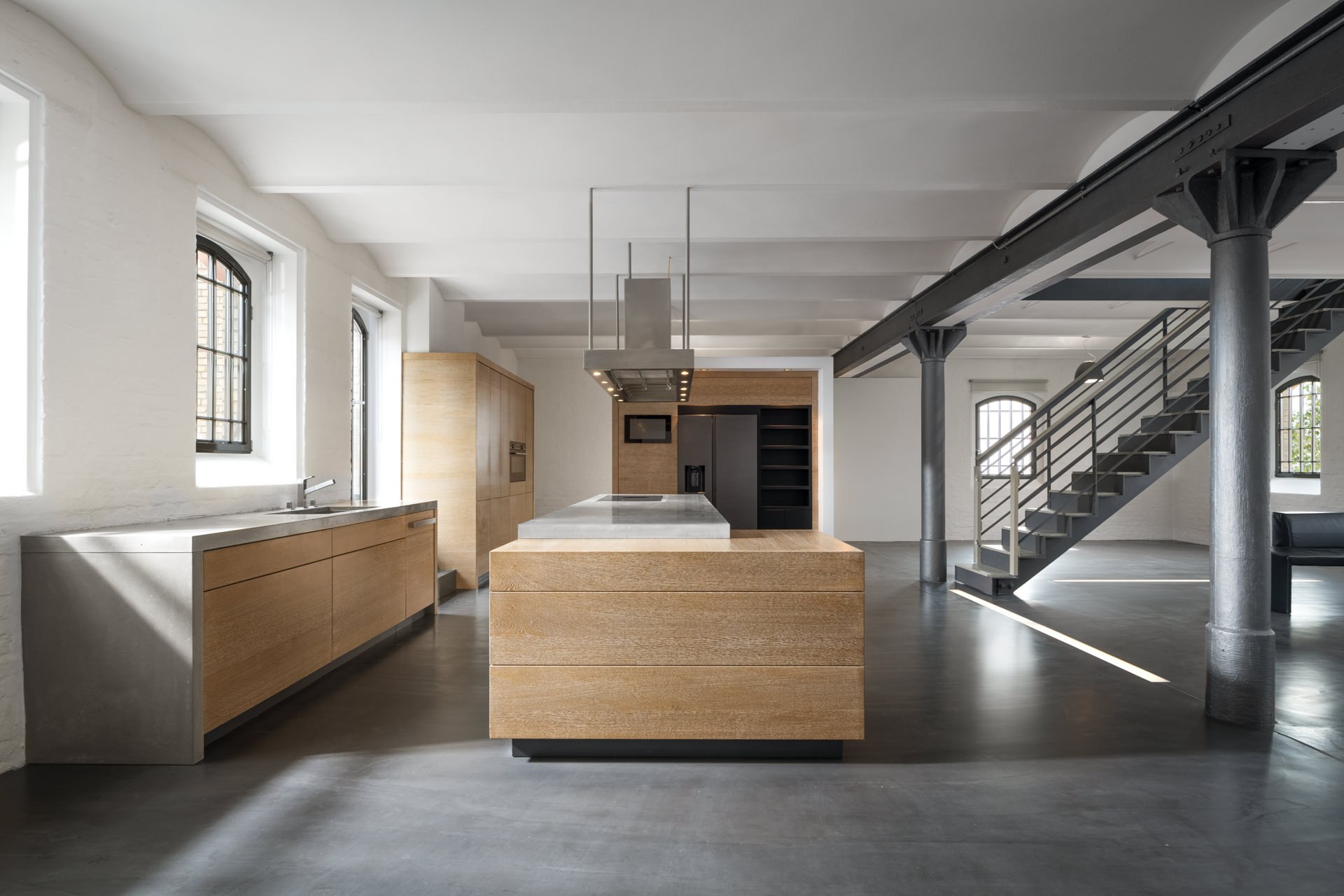Open-plan industrial loft kitchen with light wood furniture, concrete flooring, steel columns, and open staircase
