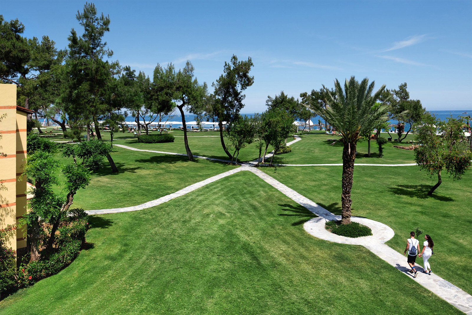 Couple walking along a winding garden path at a beachfront Mediterranean resort with lush lawns, palm trees, and the sea in the background — hospitality photography