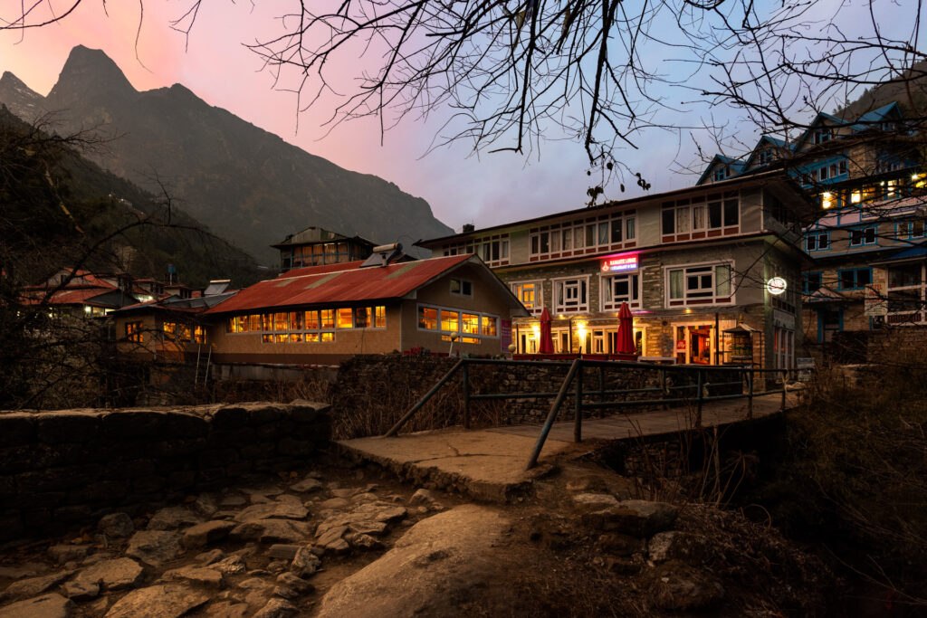 Illuminated mountain lodge in a Himalayan village at dusk with bare trees silhouetted against a pink and blue sky