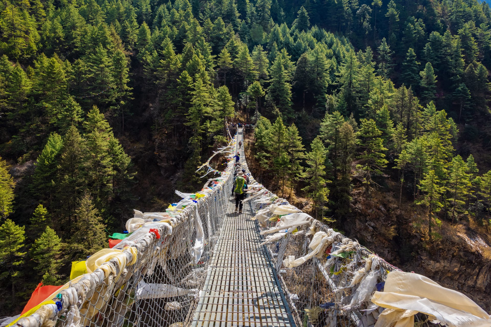 High suspension bridge adorned with colorful Tibetan prayer flags stretching over a deep forest gorge in Nepal