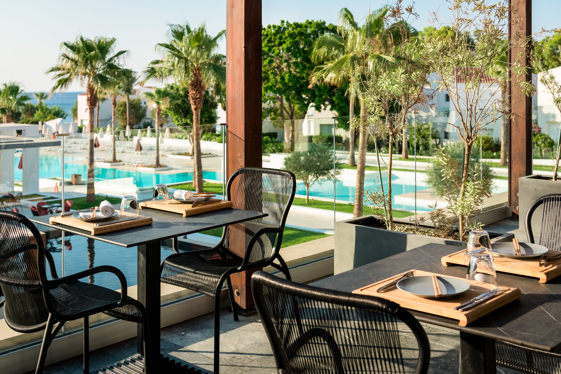 Bright outdoor restaurant terrace with rattan chairs, white tables, and a view of the resort pool and palm trees