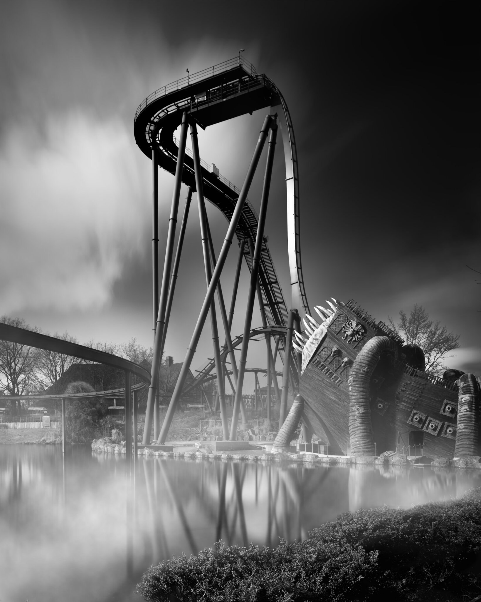 Long exposure black and white fine art photograph of a rollercoaster drop with a monster sculpture rising from morning mist