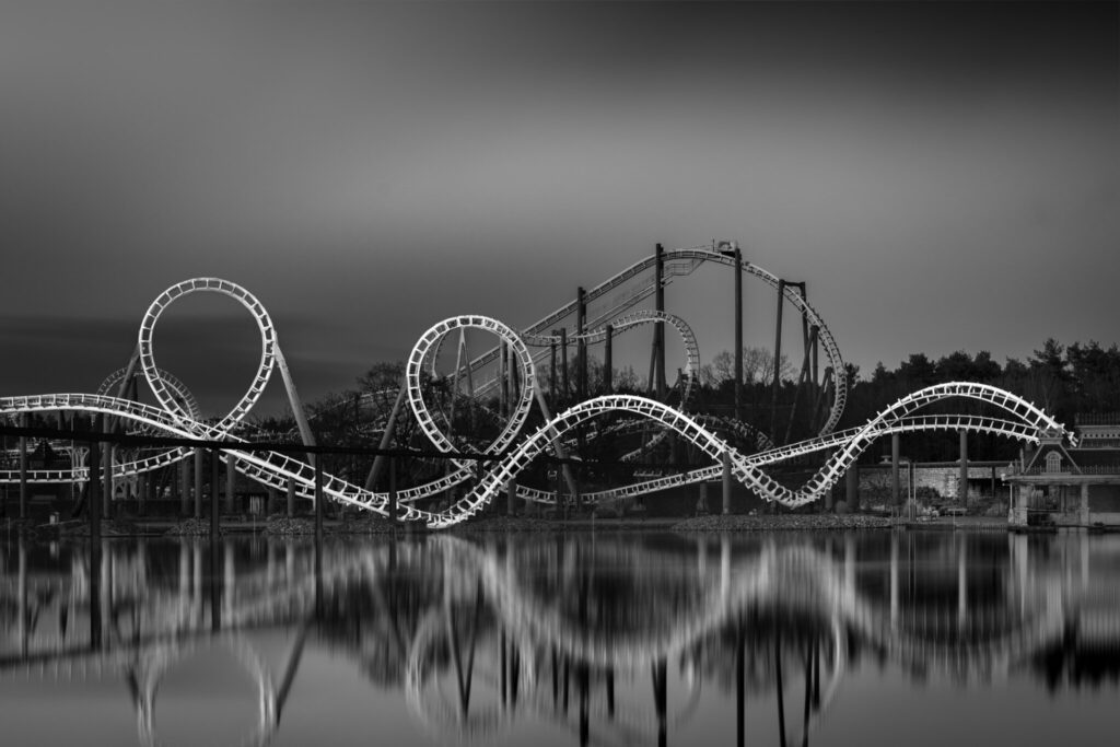 Long exposure black and white fine art photograph of rollercoaster loops perfectly reflected in a glassy lake